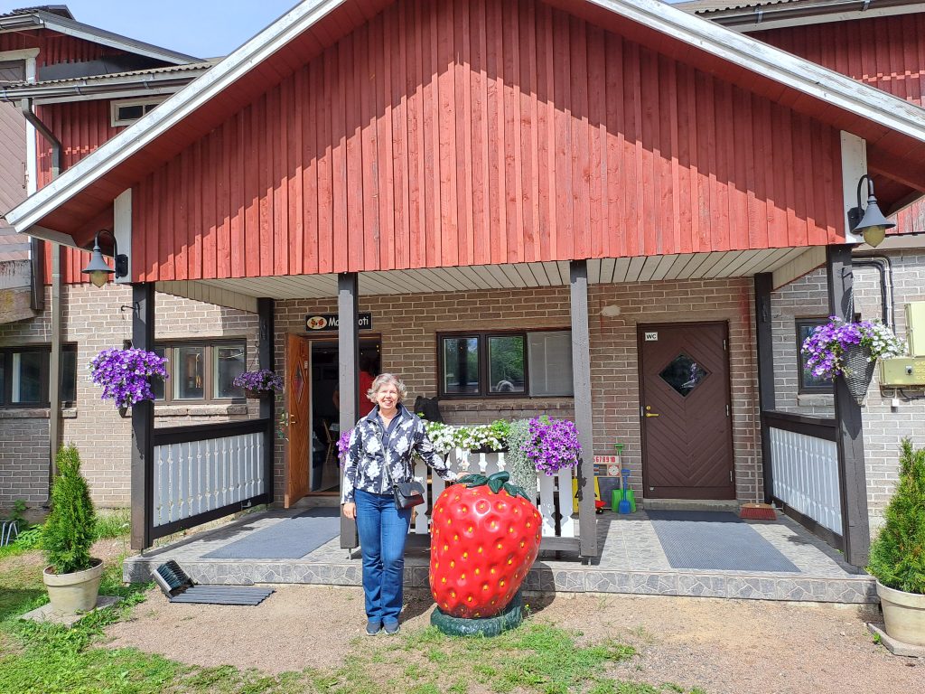 Nainen seisoo rakennuksen edessä ja nojaa isoon maassa olevaan mansikkaan.